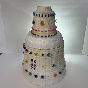 May include: A white, tiered decorative object with floral and geometric patterns. It is adorned with colourful, jewel-like accents and beaded trim in blue, pink, and yellow. The object has a bell-like shape, with a flared base and a narrower top.