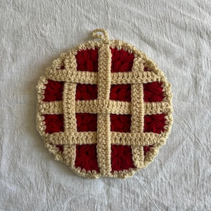 May include: A round, crocheted trivet resembling a pie. The trivet features a red filling with a beige lattice crust design. A small loop is attached for hanging. The trivet is designed to protect surfaces from hot dishes.