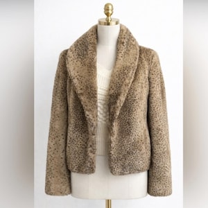 May include: A tan and brown faux fur jacket with a leopard print pattern. The jacket has a wide collar and is displayed on a mannequin. The jacket is a neutral color and can be worn with many outfits.