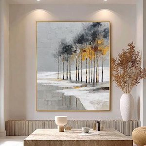 Puede incluir: Pintura abstracta de paisaje con una fila de árboles con follaje gris y dorado sobre un cielo gris. La obra de arte está enmarcada en dorado y cuelga sobre una mesa de centro de madera en un salón moderno.