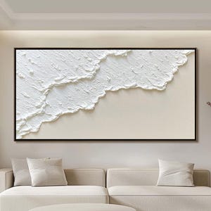 May include: A rectangular artwork in a black frame, depicting a textured white design against a beige backdrop, reminiscent of waves. The piece hangs above a beige sofa with matching cushions. The overall aesthetic is modern and minimalist, suitable for a living room.