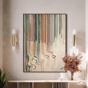 Op de afbeelding: Abstract kunstwerk met een zwarte lijst, met verticale lijnen in groen, oranje en bruin die overgaan in golvende patronen. Het kunstwerk hangt boven een gouden en houten console, met een kleine, ronde, beige poef ervoor.