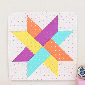 May include: A framed, colorful star quilt block with yellow, purple, orange, and teal segments. The frame is pink, and the background is a light canvas. Sewing supplies, including spools of yellow thread, a pink bear-shaped coin purse, and pink scissors, are arranged around the frame.