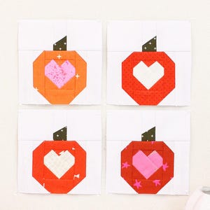May include: A framed artwork featuring four fabric blocks with pumpkin and heart designs. The pumpkins are orange and red with green stems, and the hearts are pink and white. The frame is light pink, and there are spools of yarn and a pumpkin-shaped mug in the foreground.
