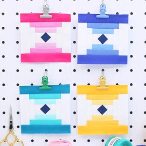 May include: Four colourful geometric art prints clipped to a white pegboard. Each print features a diamond shape and gradient colours: pink, blue, teal, and yellow. Spools of yarn and gold scissors are also visible.