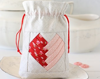 May include: A gray fabric pouch with a red drawstring and a heart-shaped patchwork design in red and pink fabrics. The pouch is sitting on a white plate with red and pink buttons.