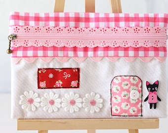May include: A pink and white gingham fabric pouch with a lace trim and a zipper. The pouch is decorated with embroidered flowers and a black cat applique. The pouch is displayed on a wooden easel.