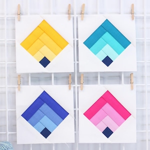 Puede incluir: Cuatro piezas de arte textil cuadradas con diseños geométricos en tonos amarillos, azules y rosas, cada una con un cuadrado azul oscuro en la parte inferior. Las piezas están sujetas a un fondo de rejilla blanca con pequeñas pinzas de madera.