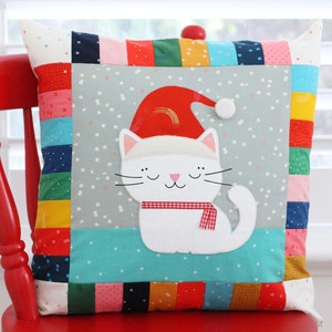 Könnte beinhalten: Ein farbenfrohes Patchwork-Kissen mit einer weißen Katze, die einen roten Weihnachtsmann-Hut und einen rot-weißen Schal trägt. Die Katze sitzt auf einem grauen Hintergrund mit weißen Schneeflocken. Das Kissen sitzt auf einem roten Stuhl.