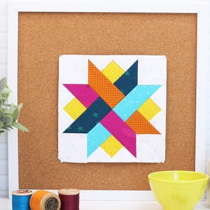 May include: A colorful star quilt block displayed on a corkboard. The star design features orange, yellow, teal, pink, and navy blue fabrics. The block is mounted in a white frame, with spools of thread and a yellow bowl in the foreground.