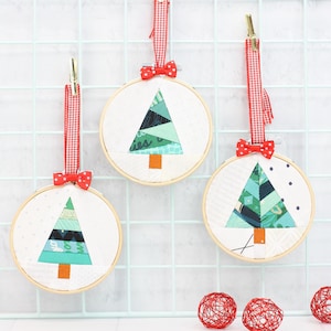 Könnte beinhalten: Drei handgemachte Weihnachtsornamente mit grünen und blauen gemusterten Papierbäumen in weißen Stickrahmen mit rot-weißem Gingham-Band und roten Schleifen.