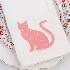 May include: White linen napkin with a pink cat silhouette and white polka dots. The napkin is folded on a white plate with a floral rim. A decorative table setting, perfect for a cat lover.