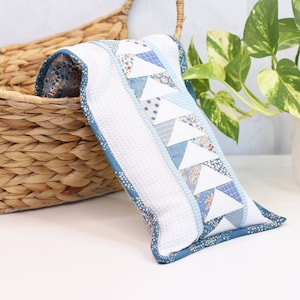 May include: A white and blue patchwork pillow with a geometric pattern of triangles. The pillow is sitting on a white surface next to a wicker basket and a potted plant.