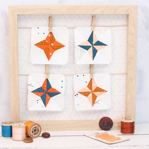 May include: Four fabric squares with a star design in shades of orange, pink, blue, and white are displayed on a wooden frame. The squares are hung with clothespins.