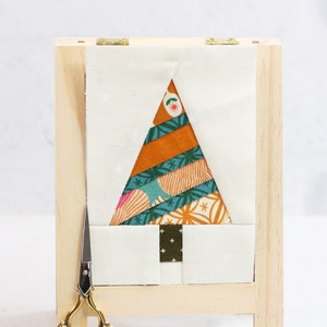 Puede incluir: Un marco de madera contiene una tela blanca con un diseño de árbol de Navidad de patchwork colorido. Un par de tijeras doradas y una lata verde con una etiqueta blanca están sobre una superficie blanca.