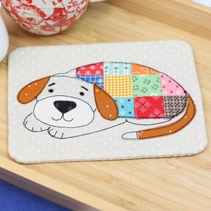 以下が含まれることがあります： カラフルなパッチワークの犬のデザインが施された白い布製のコースター。犬は足を組んで横たわっています。パッチワークは、さまざまな模様のカラフルな布の切れ端で作られています。コースターは、熱い飲み物から表面を保護するのに最適です。