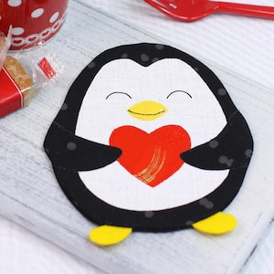 May include: A penguin-shaped coaster with a smiling face and a red heart. The coaster is black and white with yellow feet. A red fork and a red and white polka-dot cup are in the background.