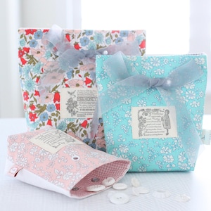 Może przedstawiać: Trzy torby na prezenty z tkaniny w kwiaty, zawiązane przezroczystymi wstążkami. Torby są w odcieniach czerwieni, niebieskiego i różu, z białymi wzorami kwiatowymi. Każda torba ma etykietę z tekstem. Białe guziki są rozrzucone przed różową torbą.