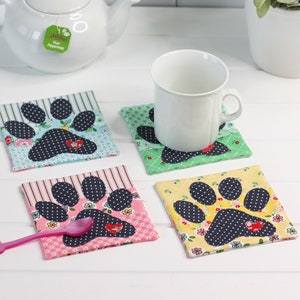 May include: Four fabric coasters with a paw print design in blue, yellow, pink, and green. The coasters are made of different patterned fabrics and feature a small red heart in the center of each paw print.