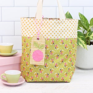 May include: A yellow and green fabric tote bag with a pink apple tag. The bag has a white polka dot pattern on the top half and a floral pattern on the bottom half. The bag has two white straps.