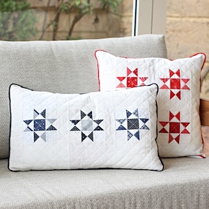 May include: Two white throw cushions with a quilted design. One cushion has a red and white star pattern, and the other has a blue and white star pattern. The cushions are on a grey sofa.