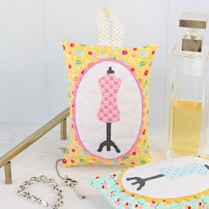May include: Two small fabric sachets with a floral pattern. Each sachet features a white oval with a black and white illustration of a dress form. One sachet is yellow with a pink dress form and the other is green with a blue dress form.