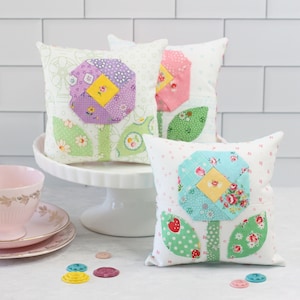 May include: Three handmade fabric flower pillows with a white background. The flowers are made from patchwork fabric in shades of pink, purple, blue, and green. The pillows are sitting on a white cake stand.