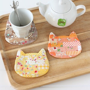 Three patchwork coasters shaped like a cat's head in colours of yellow, orange and brown,  are sitting on a wooden tray with cup, spoon and teapot.