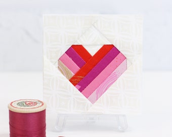 May include: A small white card with a pink and red fabric heart design. The card is on a small easel and there is a spool of pink thread and two pink buttons on a white surface.