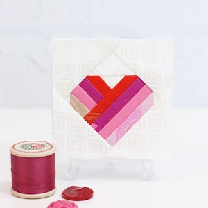 May include: A small white card with a pink and red fabric heart design. The card is on a small easel and there is a spool of pink thread and two pink buttons on a white surface.