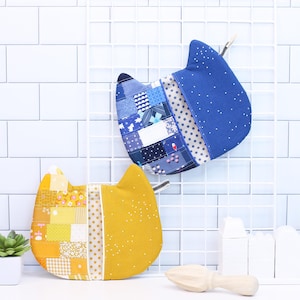 Puede incluir: Dos agarraderas para ollas con forma de gato, una azul y otra amarilla, con un diseño de patchwork. Las agarraderas están colgadas en una rejilla de alambre blanca.