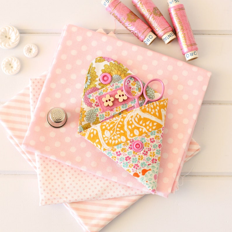 K&ouml;nnte beinhalten: Ein rosa-wei&szlig;es N&auml;hset aus Stoff mit einem gelben, rosa und gr&uuml;nen Blumendesign. Das Set enth&auml;lt eine Schere und einen Fingerhut.