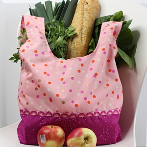 May include: A pink reusable shopping bag with a white polka dot pattern and a pink and purple trim. The bag is filled with groceries, including a loaf of bread, leeks, and leafy greens. Two red apples are sitting on a white surface in front of the bag.