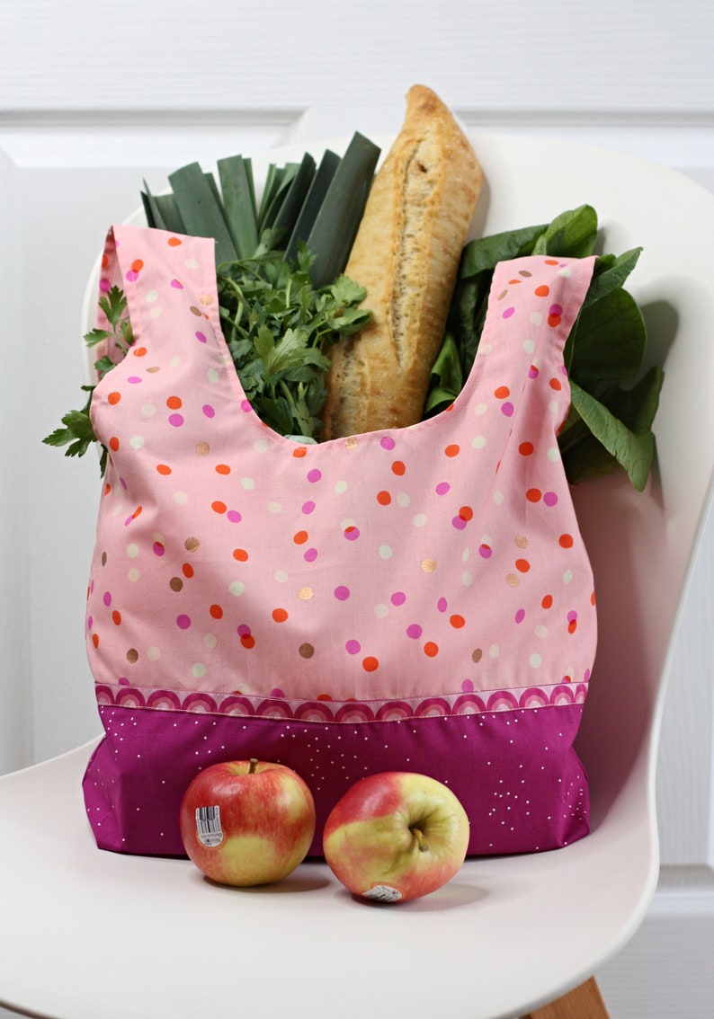 May include: A pink reusable shopping bag with a white polka dot pattern and a pink and purple trim. The bag is filled with groceries, including a loaf of bread, leeks, and leafy greens. Two red apples are sitting on a white surface in front of the bag.
