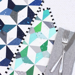 Puede incluir: Dos manteles individuales acolchados con un patrón geométrico en tonos azules, verdes y blancos. Los manteles individuales tienen un ribete blanco y negro. Una servilleta gris con un tenedor y un cuchillo plateados está encima de los manteles individuales.