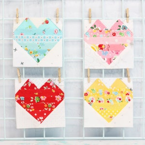 May include: Four fabric heart blocks, each with a different color scheme. The blocks are displayed on a white grid background. The hearts are made with a patchwork of different fabrics, including floral prints, polka dots, and stripes. The colors of the hearts are red, pink, yellow, and blue.