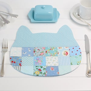 Peut inclure: Un set de table en forme de chat en patchwork bleu et blanc avec une bordure bleu clair. Le set de table est fait de différents tissus avec des motifs floraux et géométriques.