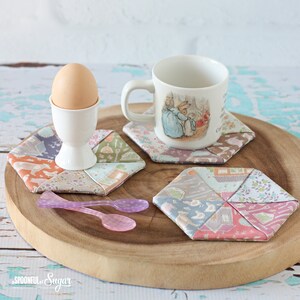 May include: A wooden serving tray holds a white mug with a bunny illustration, an egg in a white egg cup, and three hexagonal fabric coasters. Two small pink spoons are also on the tray. The coasters are made of patchwork fabric in pastel colors.