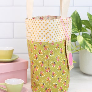 K&ouml;nnte beinhalten: Eine gelbe und gr&uuml;ne Stofftasche mit wei&szlig;en Punkten und Blumendruck. Die Tasche hat zwei wei&szlig;e Stoffgriffe und ein rosa Stoff-Etikett mit wei&szlig;em Punktmuster.