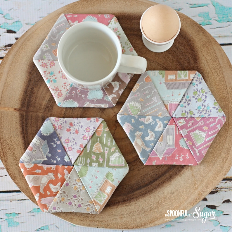 May include: A white ceramic mug sits on a patchwork hexagon coaster. Two additional coasters and a soft-boiled egg in a white egg cup are also visible. The coasters are made of fabric with floral and pastoral patterns in pastel colors.