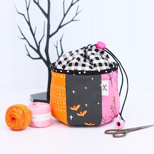 Pode incluir: Uma bolsa com cord&atilde;o de patchwork com uma parte superior xadrez em preto e branco, pain&eacute;is laranja, cinza e rosa. O painel cinza apresenta morcegos e estrelas. Dois carret&eacute;is de linha e tesouras est&atilde;o ao lado da bolsa. Uma &aacute;rvore decorativa preta est&aacute; no fundo.