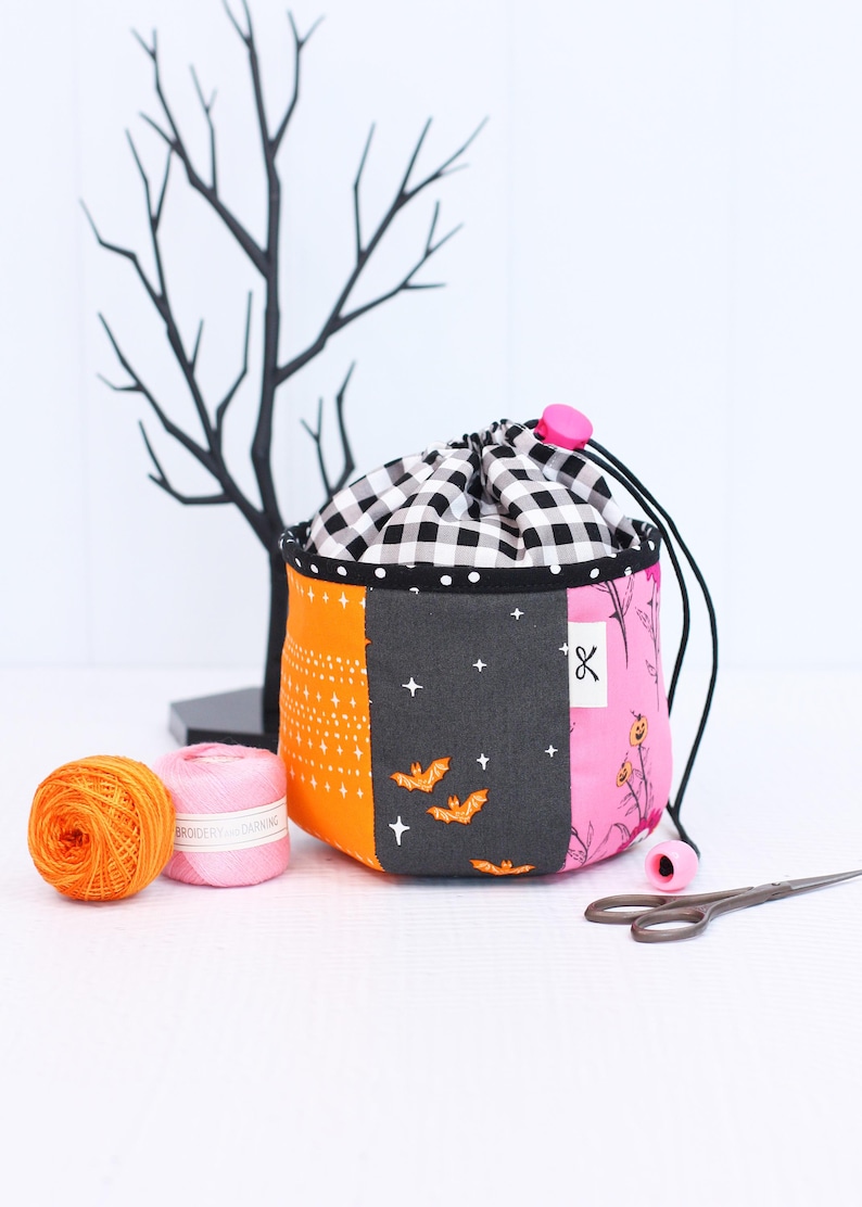 Pode incluir: Uma bolsa com cord&atilde;o de patchwork com uma parte superior xadrez em preto e branco, pain&eacute;is laranja, cinza e rosa. O painel cinza apresenta morcegos e estrelas. Dois carret&eacute;is de linha e tesouras est&atilde;o ao lado da bolsa. Uma &aacute;rvore decorativa preta est&aacute; no fundo.