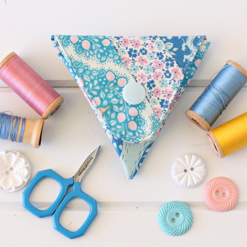 K&ouml;nnte beinhalten: Eine dreieckige N&auml;hbox aus Stoff mit einem blauen und rosa floralen Muster. Die Box hat einen Knopfverschluss und ist mit N&auml;hzubeh&ouml;r gef&uuml;llt. Die Box steht auf einer wei&szlig;en Holzoberfl&auml;che mit anderem N&auml;hzubeh&ouml;r, darunter Garnrollen, Kn&ouml;pfe und eine kleine Schere.