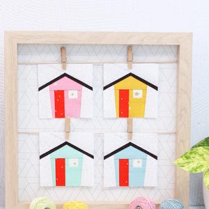 May include: Four small fabric squares with a house design in pink, yellow, blue, and green. Each house has a red door and a white window with a gold star. The squares are displayed in a wooden frame with clothespins.