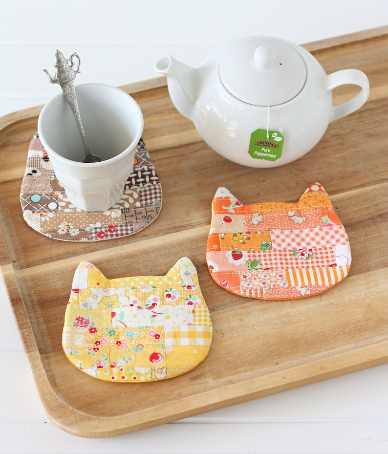 Three patchwork coasters shaped like a cat's head in colours of yellow, orange and brown,  are sitting on a wooden tray with cup, spoon and teapot.