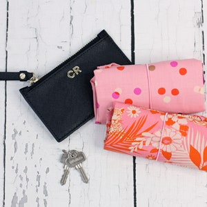 May include: A black leather zippered pouch with the initials "CR" embossed on the front. The pouch is next to two pink fabric bags with floral and polka dot patterns. The bags are tied with pink elastic bands.