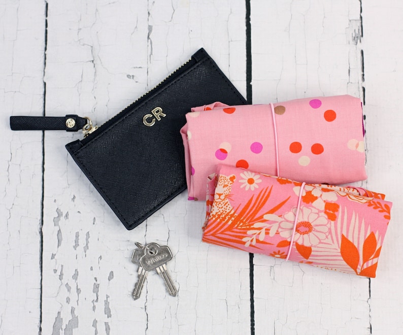 May include: A black leather zippered pouch with the initials "CR" embossed on the front. The pouch is next to two pink fabric bags with floral and polka dot patterns. The bags are tied with pink elastic bands.