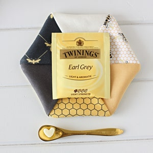 May include: A hexagonal fabric coaster with a tea bag and a small spoon. The coaster is made of various fabrics, including a black and gold bee pattern, a honeycomb pattern, and solid colors. The tea bag is labeled "Twinings Earl Grey".