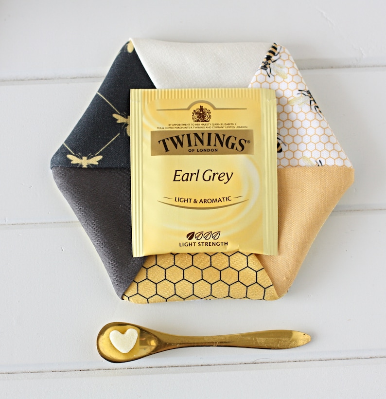 May include: A hexagonal fabric coaster with a tea bag and a small spoon. The coaster is made of various fabrics, including a black and gold bee pattern, a honeycomb pattern, and solid colors. The tea bag is labeled "Twinings Earl Grey".