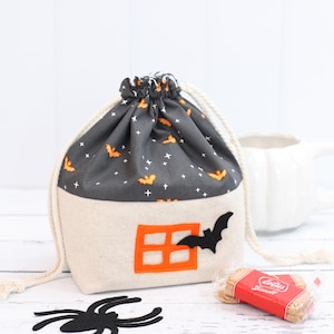 May include: A gray and white Halloween-themed drawstring bag with a bat and window design. The bag is filled with cookies and a black spider is on the table.